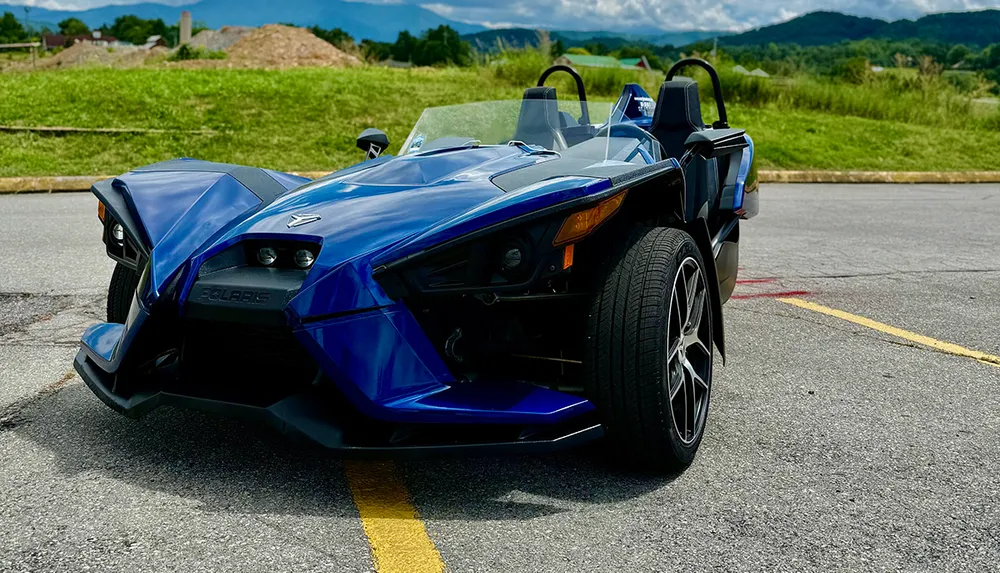 A striking blue three-wheeled vehicle with angular design features is parked in an outdoor setting with mountains in the background