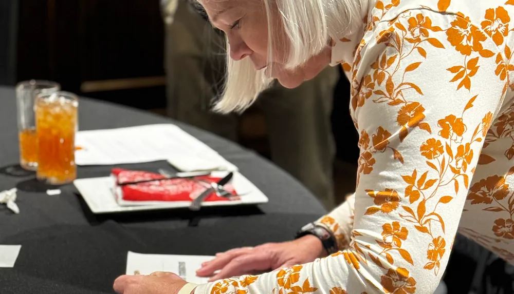 A person wearing a floral-patterned shirt is leaning over a table writing on a piece of paper with drinks and a plate nearby