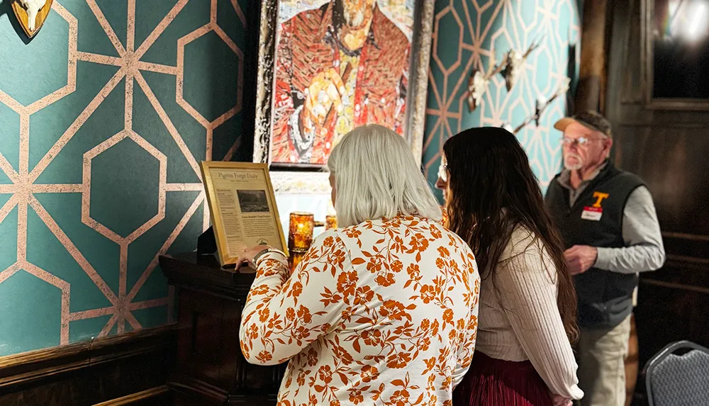 Three people are observing a framed newspaper article and a colorful portrait in a room with a decorative green and pink geometric patterned wall