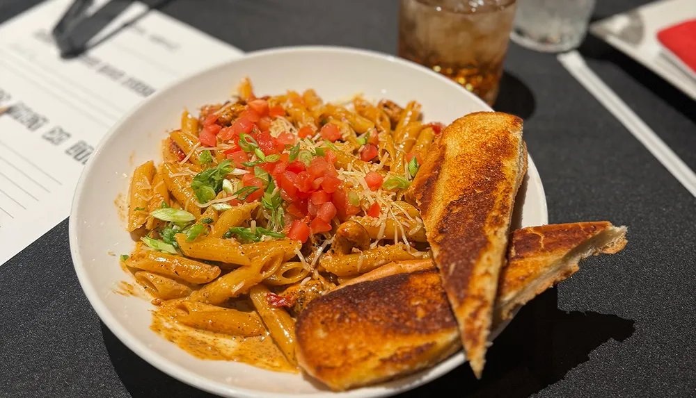A plate of penne pasta topped with diced tomatoes and herbs is served alongside toasted bread with a drink in the background