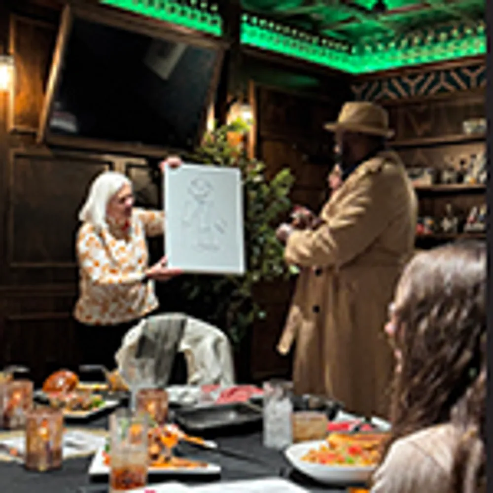 A group of people gathered around a table are playing Pictionary with one person holding up a whiteboard showing drawings
