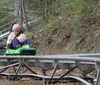 A man and a child are joyfully riding a green mountain coaster on a track surrounded by rocks and greenery