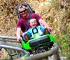 A man and a child are joyfully riding a green mountain coaster on a track surrounded by rocks and greenery