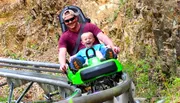 A man and a child are joyfully riding a green mountain coaster on a track surrounded by rocks and greenery.