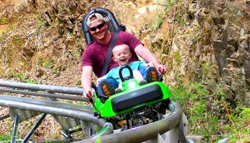 A man and a child are joyfully riding a green mountain coaster on a track surrounded by rocks and greenery.