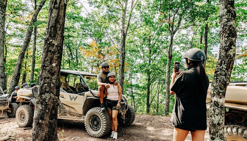 A person is taking a photo of two people posing in front of an off-road vehicle in a forested area
