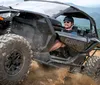 A person is enthusiastically driving a Can-Am off-road vehicle up a dirt hill with a scenic mountain view in the background