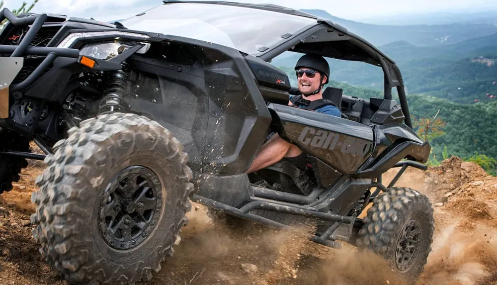 A person is enthusiastically driving a Can-Am off-road vehicle up a dirt hill with a scenic mountain view in the background