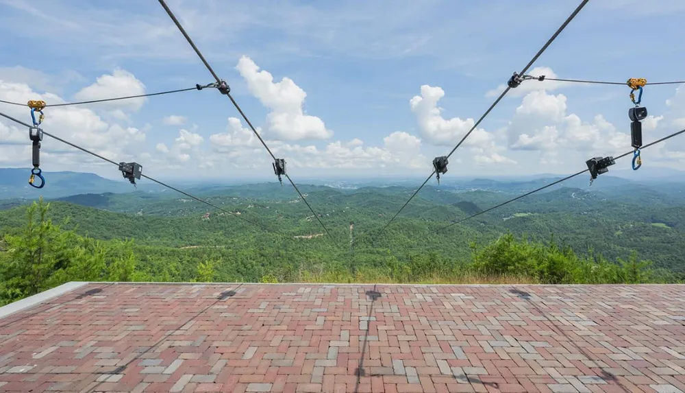 The image shows a zip line platform overlooking a vast green hilly landscape under a partly cloudy sky