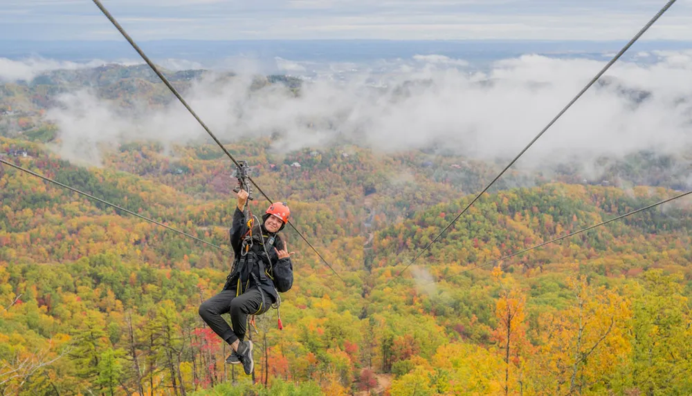 A person is zip-lining through a scenic colorful autumn forest with clouds in the background