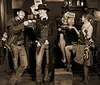 A group of people dressed in vintage Wild West attire are gathered in a saloon setting engaged in activities like card playing and socializing