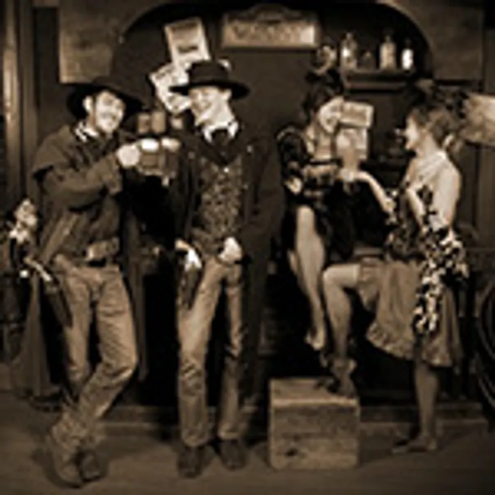 The image is a sepia-toned photograph of four people dressed in Old West-style costumes posing in a saloon setting
