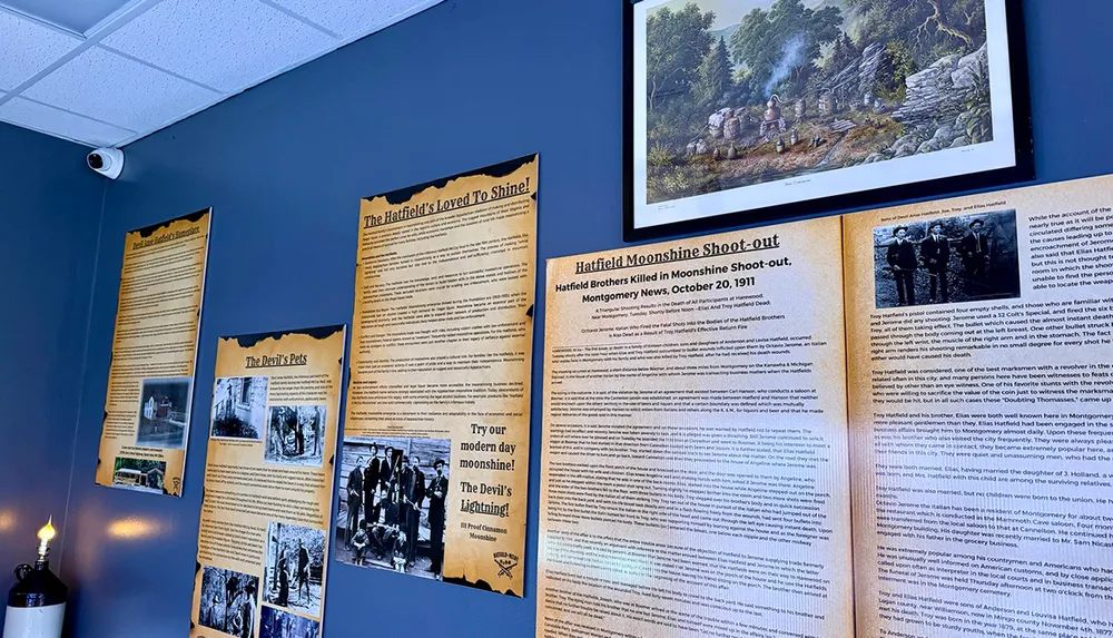 The image shows a display of historical posters and a framed painting on a wall detailing the Hatfield familys involvement with moonshine production and a notable shoot-out