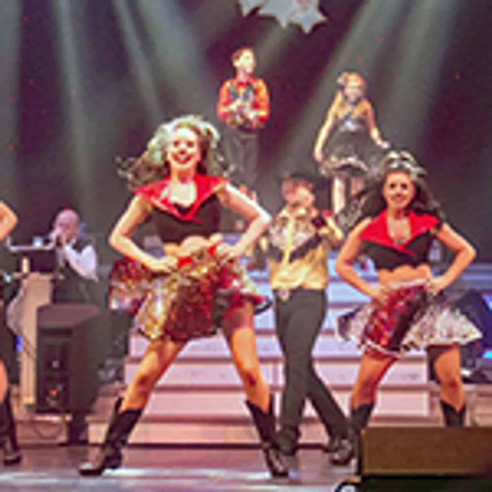 The image shows a lively stage performance with dancers wearing colorful costumes under bright spotlights