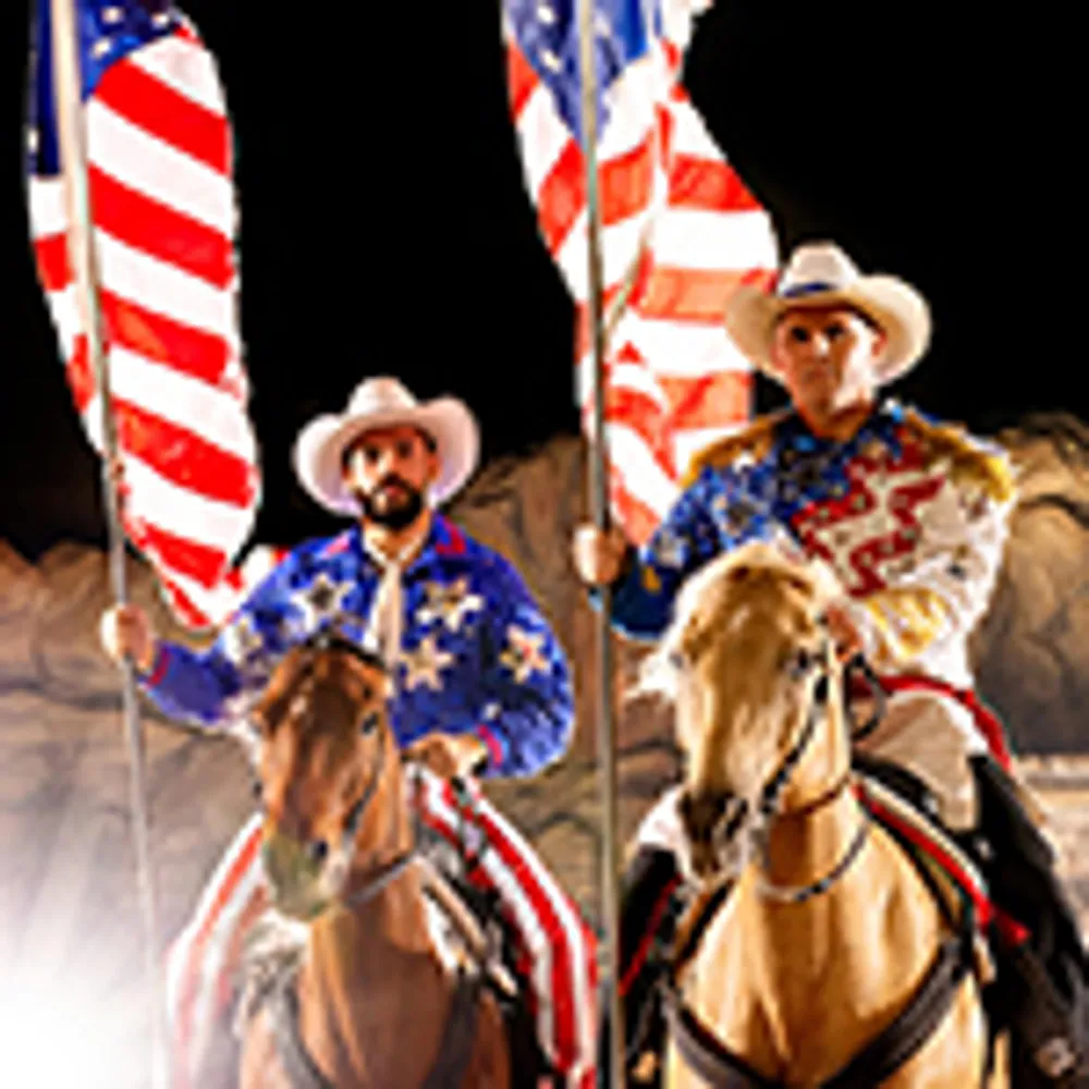 Two cowboys wearing star-spangled outfits and hats are riding horses while holding American flags