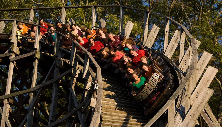 Thrilled passengers on a wooden roller coaster are captured mid-ride as they descend a steep track amidst trees.