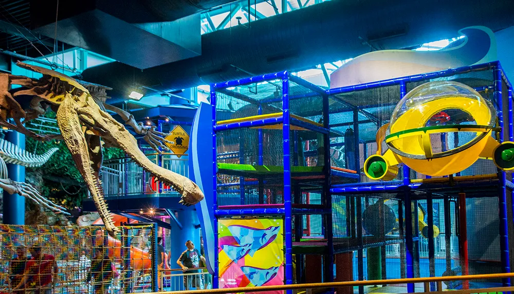 The image shows a colorful indoor playground with a multi-level climbing structure and a dinosaur skeleton exhibit creating a vibrant and educational space for children