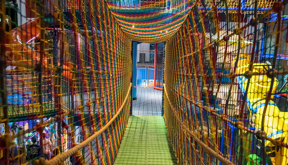 The image displays a colorful indoor playground structure with mesh tunnels and children playing inside