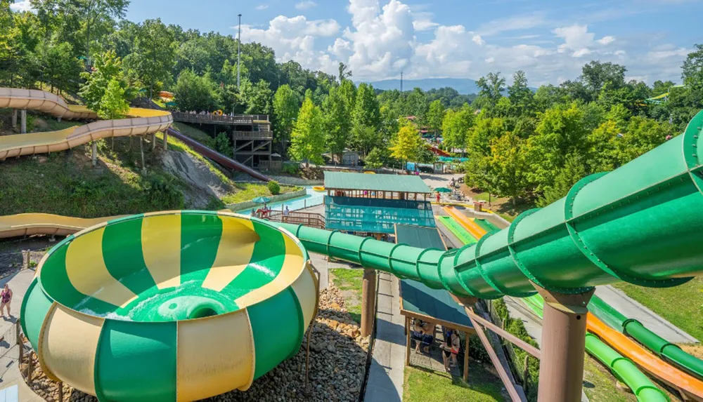The image shows a vibrant water park with various colorful slides and pools nestled in a lush green hilly landscape under a partly cloudy sky