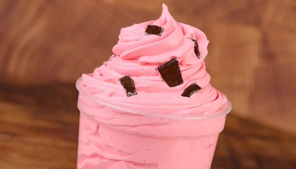 The image shows a swirl of pink soft serve ice cream topped with chocolate pieces in a clear plastic cup