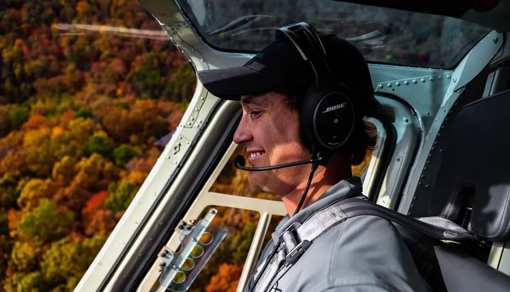 The image shows a smiling pilot wearing a headset in the cockpit of a helicopter with a backdrop of colorful autumn trees visible through the open window