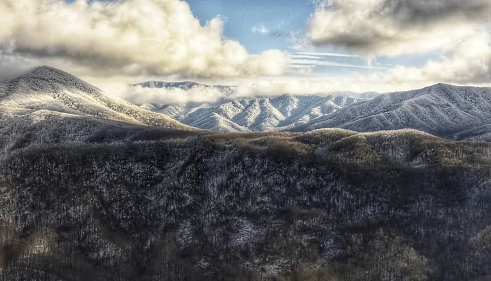 A panoramic view of snow-covered mountains under a cloudy sky with sunlight piercing through creating a dramatic landscape