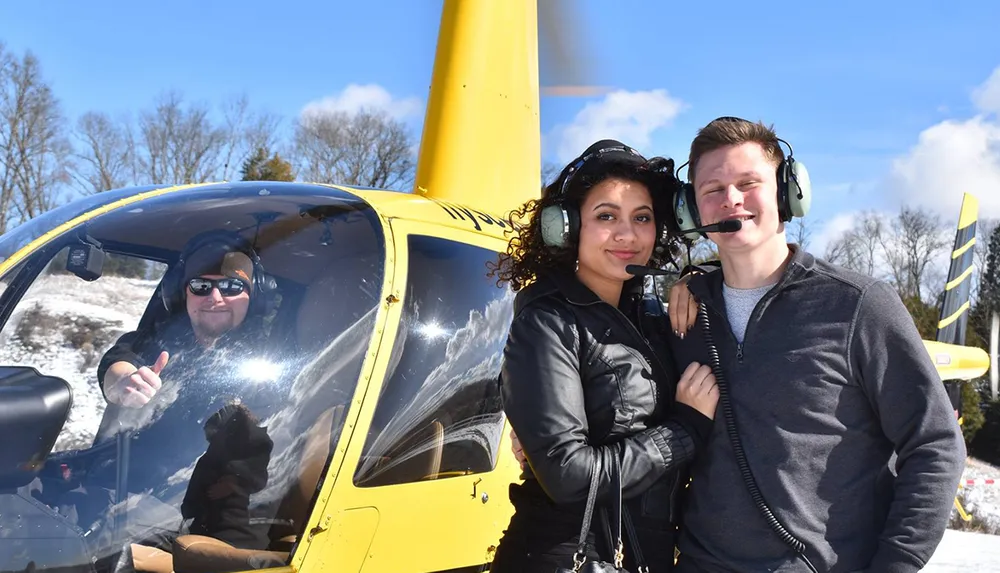 A couple stands smiling in front of a yellow helicopter wearing communication headsets while the pilot inside gives a thumbs-up