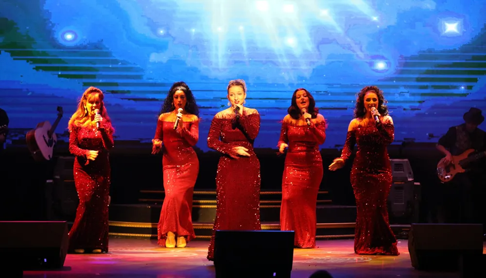 Five singers in sparkling red dresses are performing on stage with a large digital screen in the background and a musician on the right