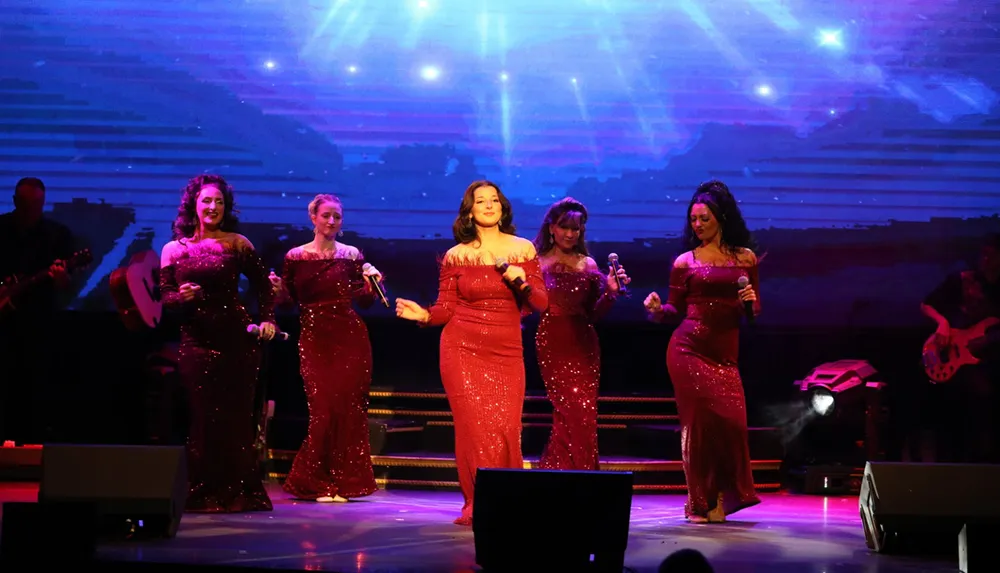 Five performers in sparkling red dresses are singing on stage with a live band in the background under colorful stage lighting