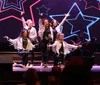 A group of performers in sparkling outfits are enthusiastically singing and dancing on a stage adorned with neon star shapes with musicians playing in the background