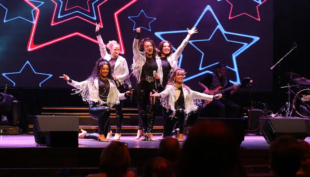 A group of performers in sparkling outfits are enthusiastically singing and dancing on a stage adorned with neon star shapes with musicians playing in the background
