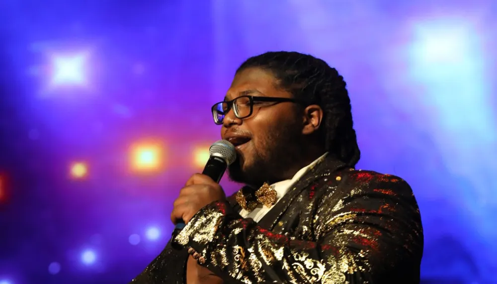 A person is singing into a microphone on a stage with a colorful bokeh light-filled background