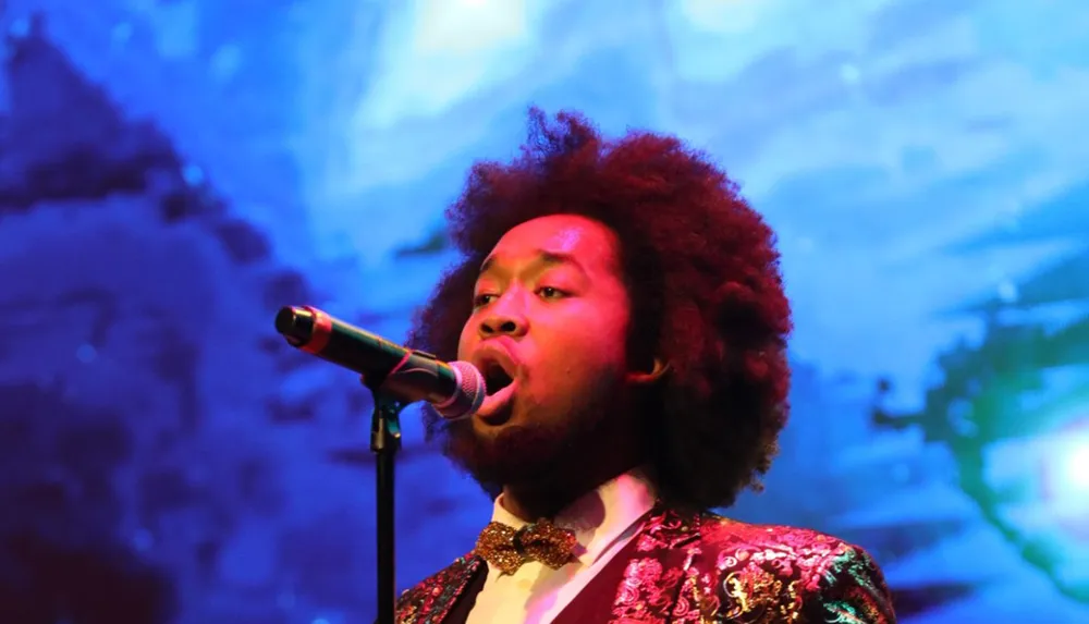 A man with a voluminous afro hairstyle is singing passionately into a microphone on a stage with vibrant blue lighting in the background