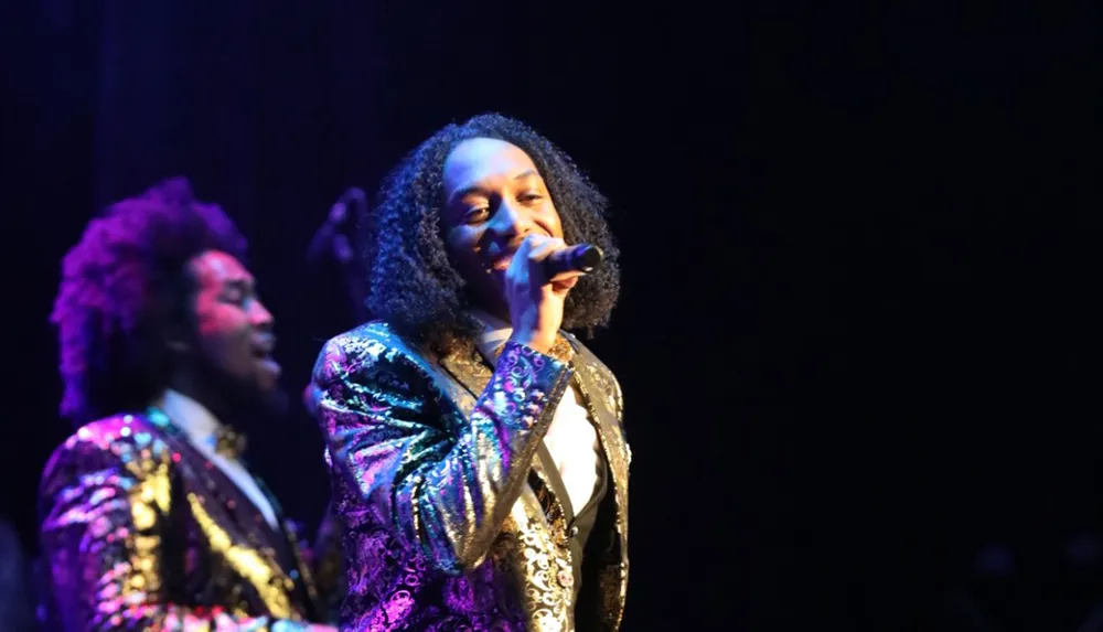 Two performers wearing sequined outfits are on stage with one singing into a microphone and the other in the background