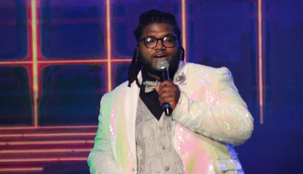 A person in a shiny suit and bow tie is singing or speaking into a microphone on stage