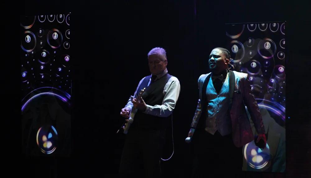 Two musicians are performing on stage with one playing a guitar and the other holding a microphone set against a backdrop with circular patterns
