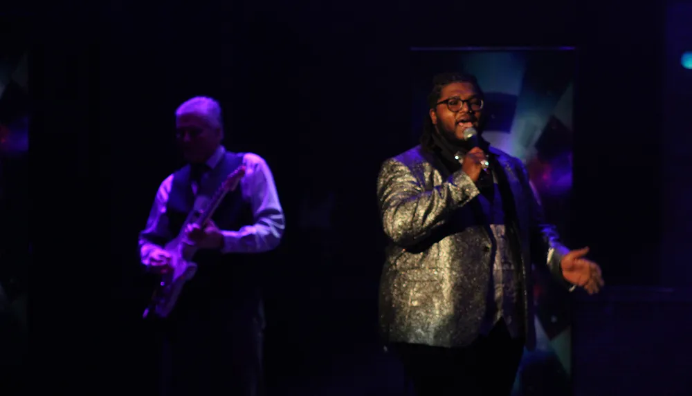 A performer in a glittery suit sings into a microphone on a dimly lit stage accompanied by a guitarist