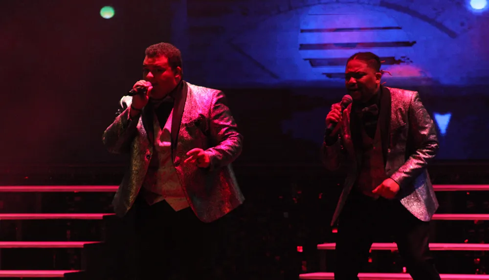 Two performers in shiny suits are singing on stage with dramatic lighting