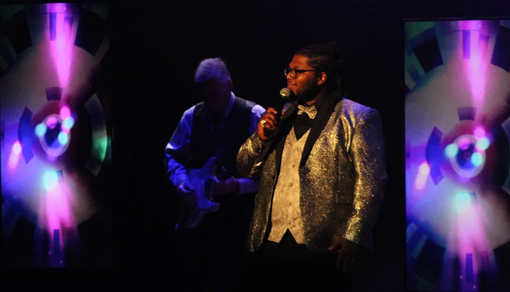 A person in a sparkly jacket sings on stage with a guitarist in the background and colorful lights on display