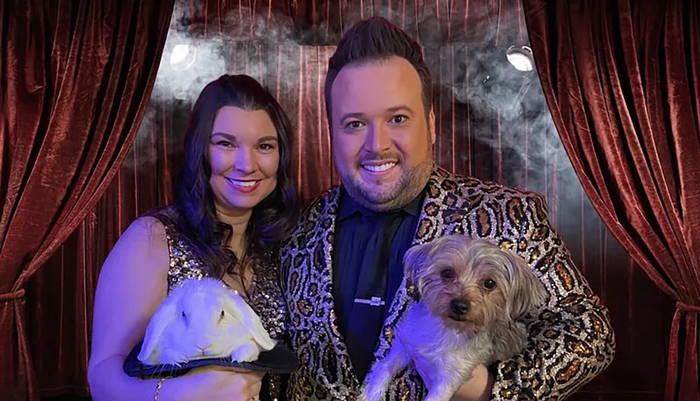A smiling man and woman in glittery attire pose behind a smoky red curtain with a white rabbit and a small dog
