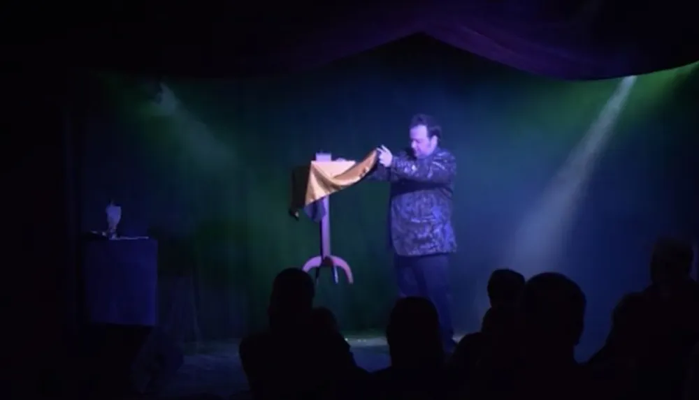 A magician is performing on stage in front of an audience with a levitated tablecloth as part of his act
