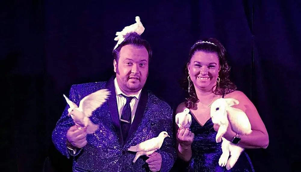A man and a woman in sparkly outfits are smiling on stage with doves in their hands and a rabbit creating a classic magic show scene