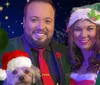 A couple dressed in festive holiday attire poses with a small dog wearing a Santa hat set against a starry night sky with a hanging green ornament