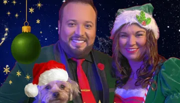 A couple dressed in festive holiday attire poses with a small dog wearing a Santa hat, set against a starry night sky with a hanging green ornament.