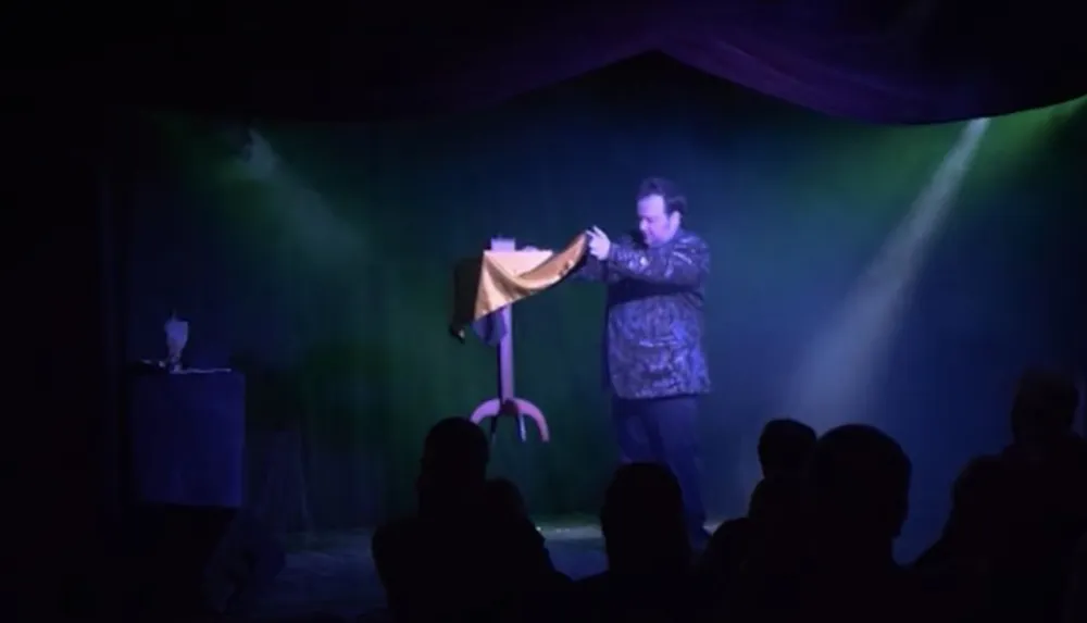 A magician is performing on stage in front of an audience with a levitated tablecloth as part of his act