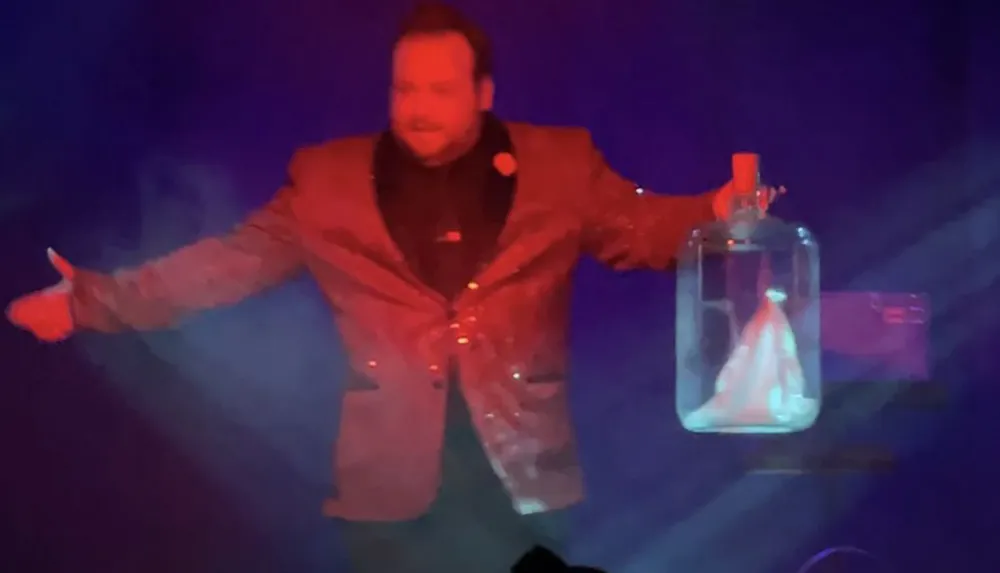 A person in a sparkly jacket performs on stage holding a glass jar with smoke inside set against colorful lighting