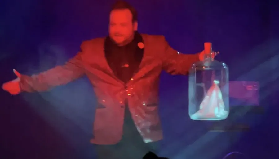 A person in a sparkly jacket performs on stage, holding a glass jar with smoke inside, set against colorful lighting.