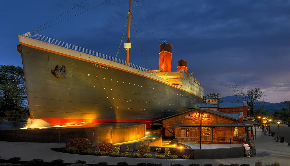The image depicts a large ship-shaped structure with the name TITANIC on the bow illuminated in the evening suggesting it is a museum or attraction rather than an actual sea-going vessel