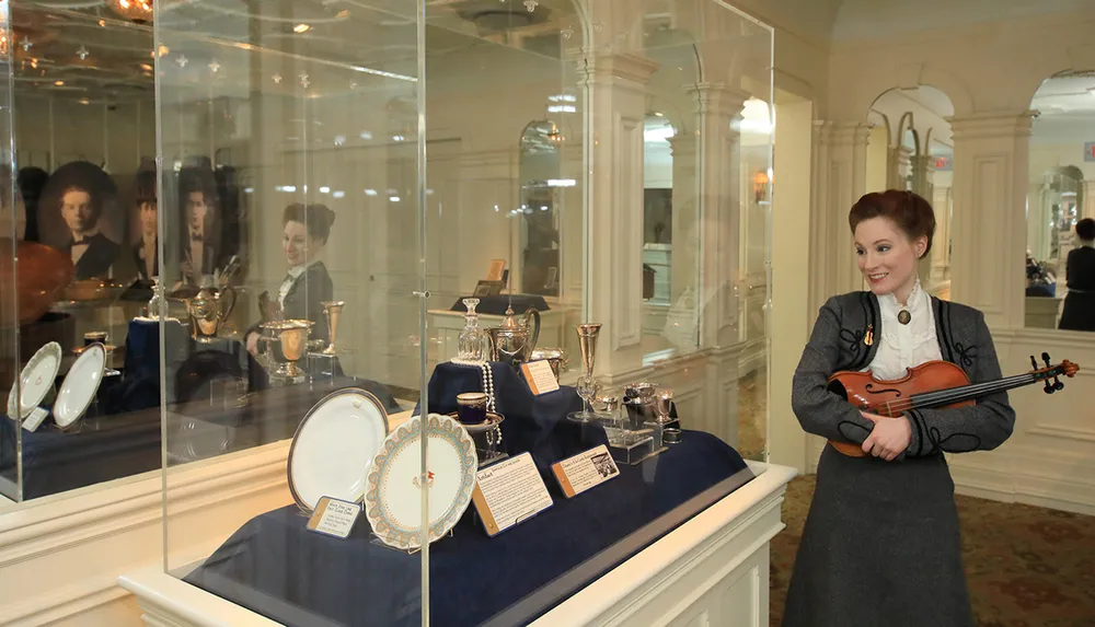 A woman holding a violin stands next to a display case containing various antique items possibly in a museum or historical exhibition