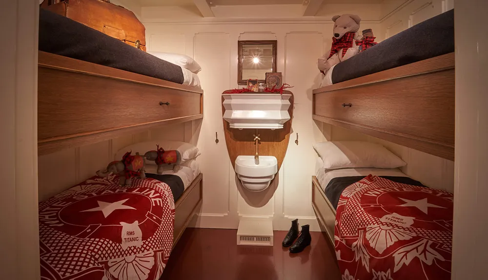 A cozy cabin features two sets of wooden bunk beds with red RMS Titanic blankets and toys a small sink and a mirror on the wall